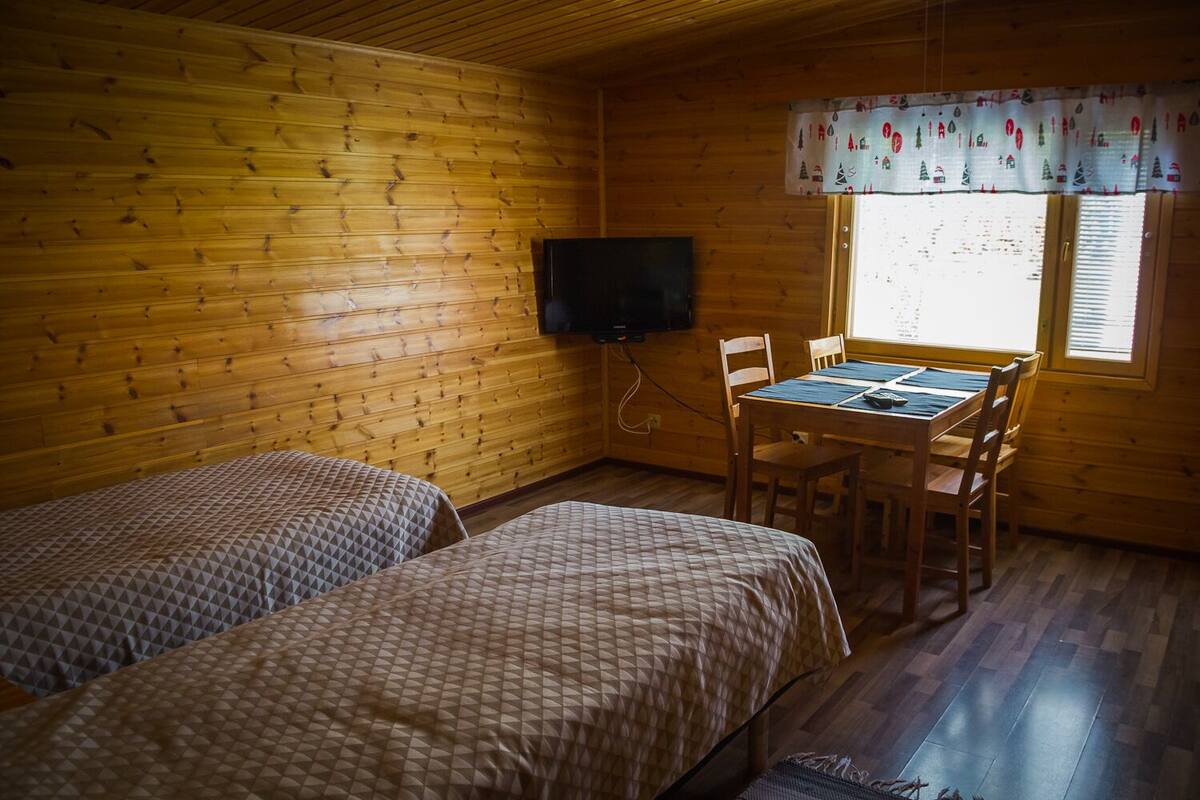 A cabin with two beds, a dining table, chairs, a television, and a window with blinds.