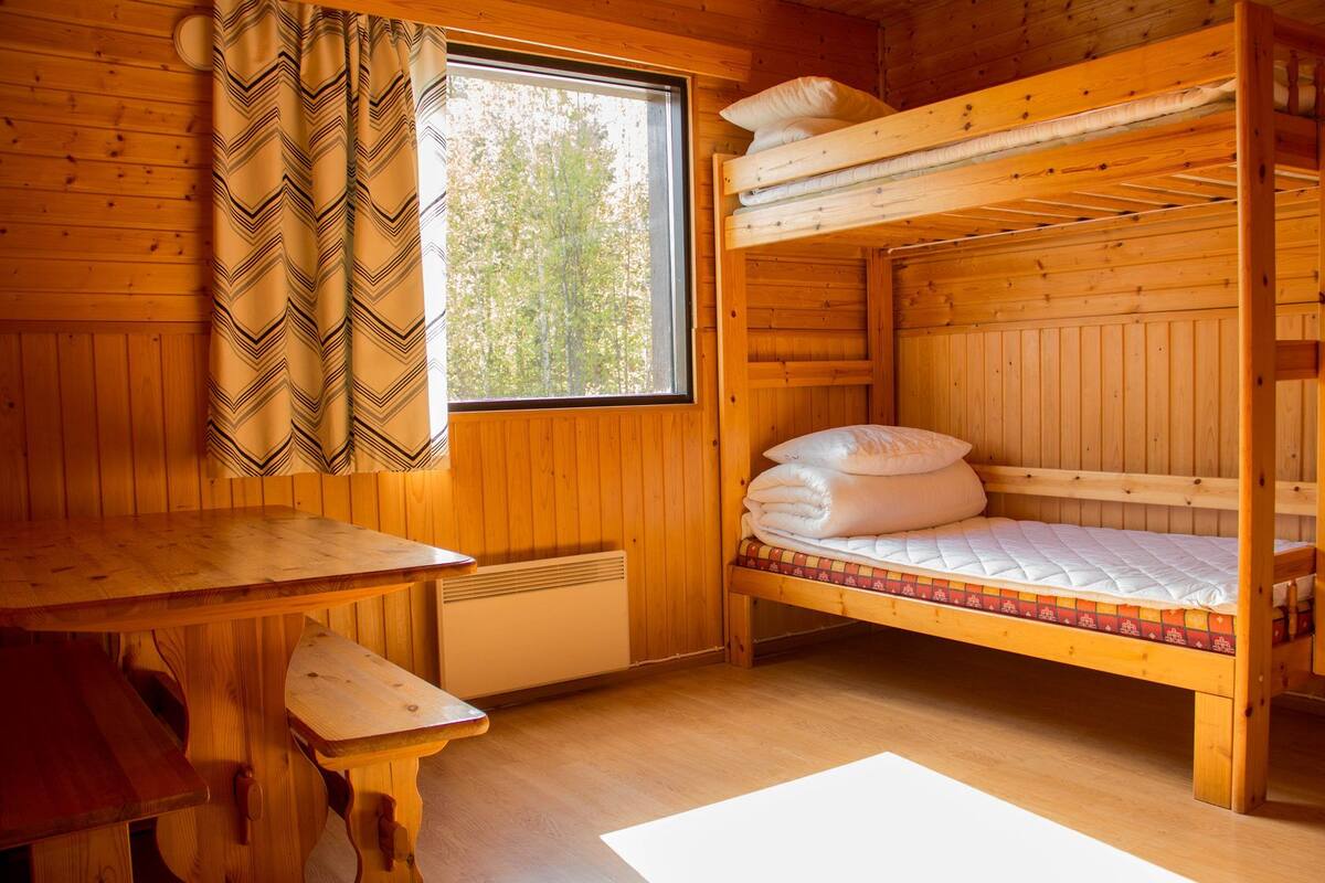 A wooden cabin room with a bunk bed, a table, a window with curtains, and a radiator.