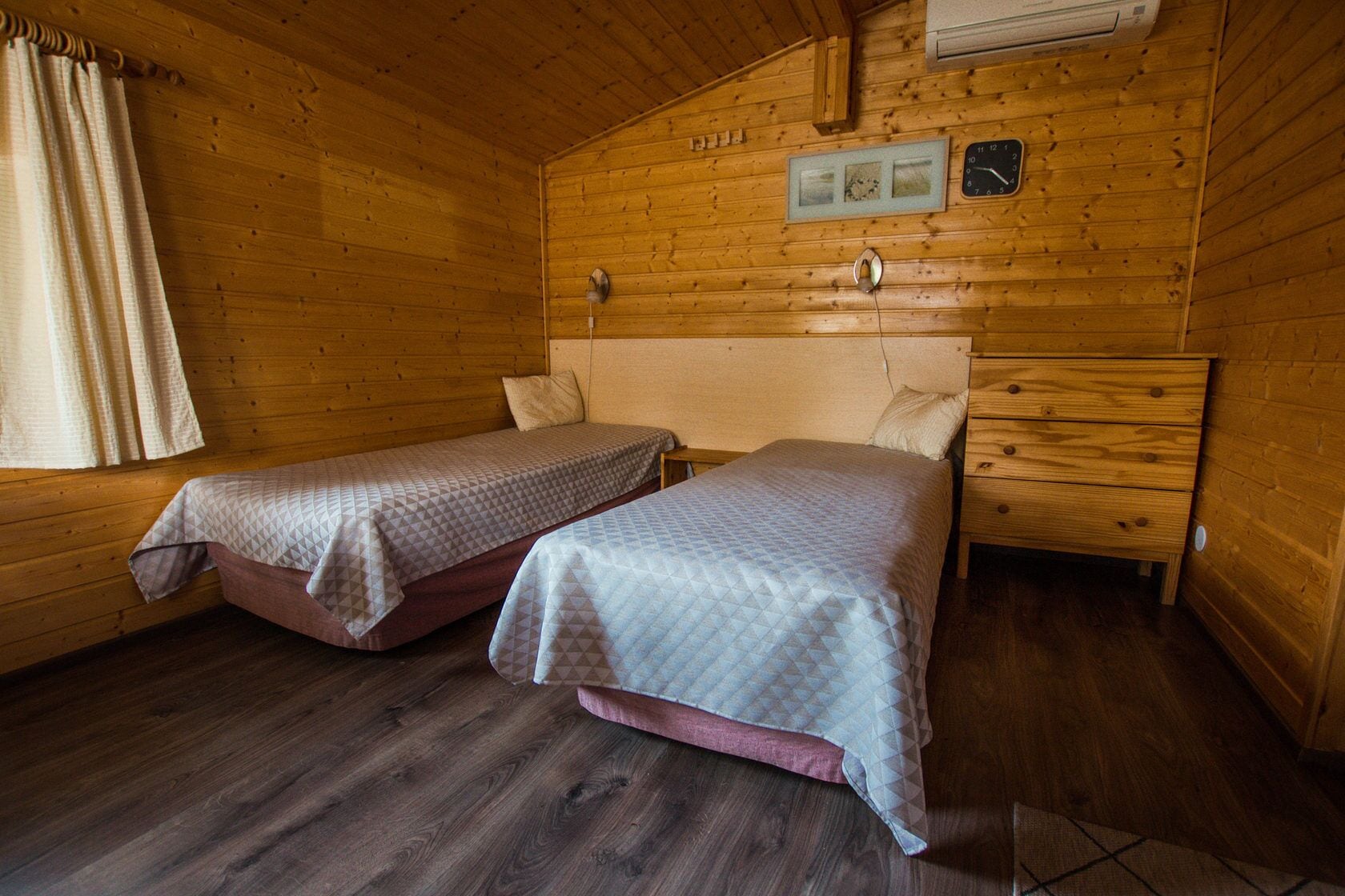A cabin-style room with two beds, wooden walls, a clock, and a small window with curtains.