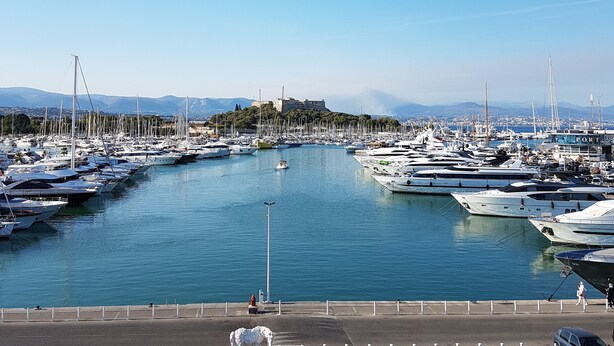 Un port de plaisance où de nombreux bateaux sont amarrés, un ciel dégagé et des montagnes à l’horizon.