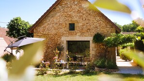A stone building with a triangular roof, a small window, and a door. There is a patio with a table and chairs, and a covered area with an umbrella.