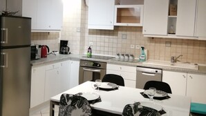 Una cocina moderna con armarios blancos, un refrigerador de acero inoxidable, un microondas y una cafetera. Hay una mesa de comedor con sillas y una ventana encima.