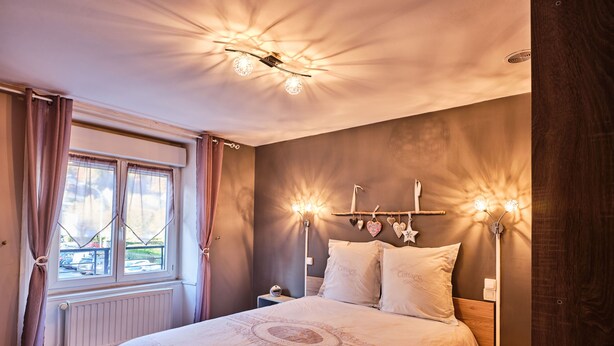 Un dormitorio con una cama grande, una mesita de noche, cortinas en una ventana e iluminación en la pared.