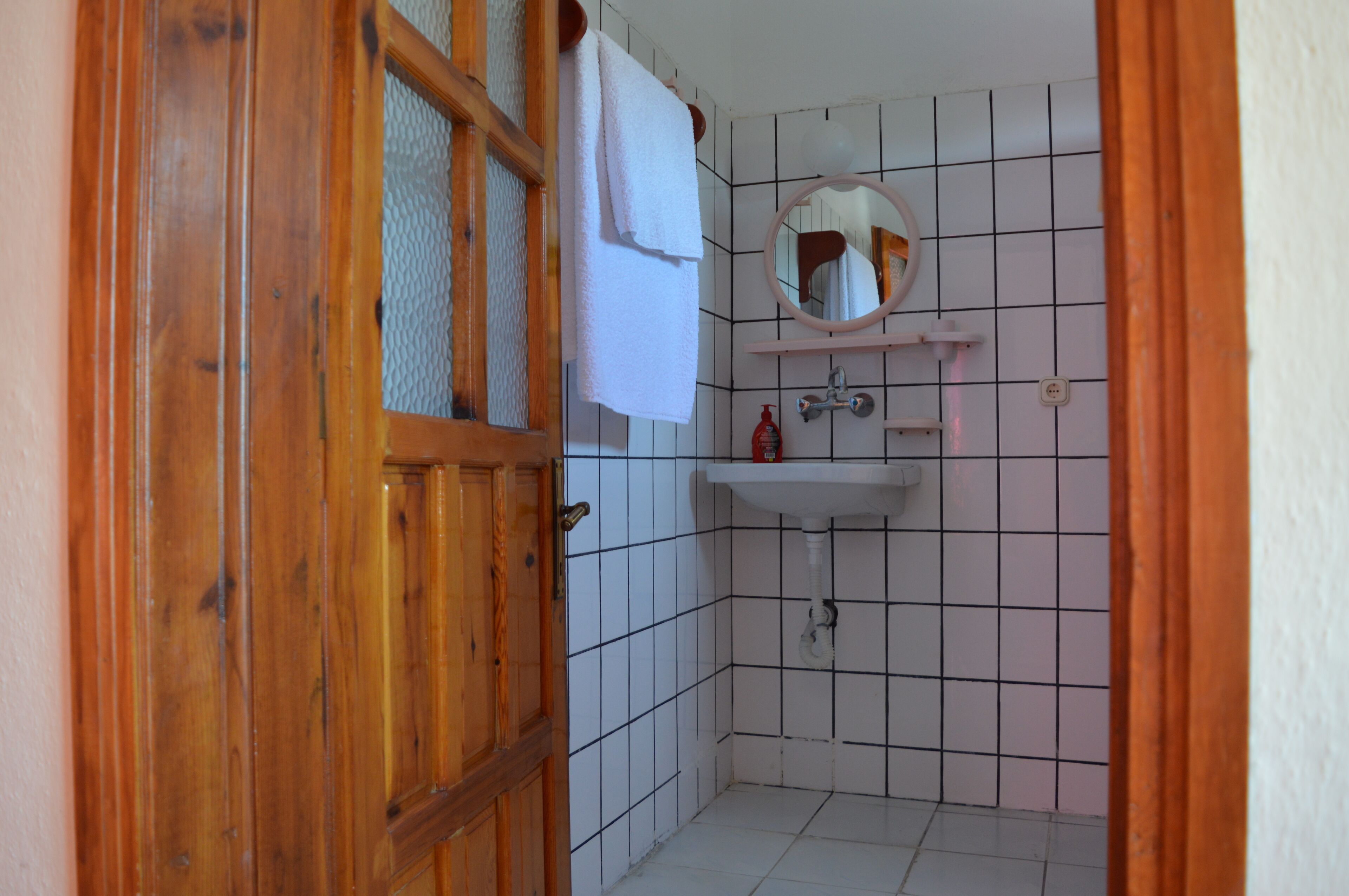 A bathroom with a wooden door, a white tiled wall, a sink with a mirror, and a towel hanging on the door.