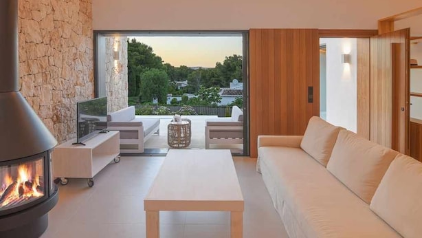 A modern living room with a stone wall, a fireplace, a sofa, and a view of a garden and buildings. - Casa Bio Nueva: Super sea views, heated pool, 100MB wifi, A / C (Jávea)