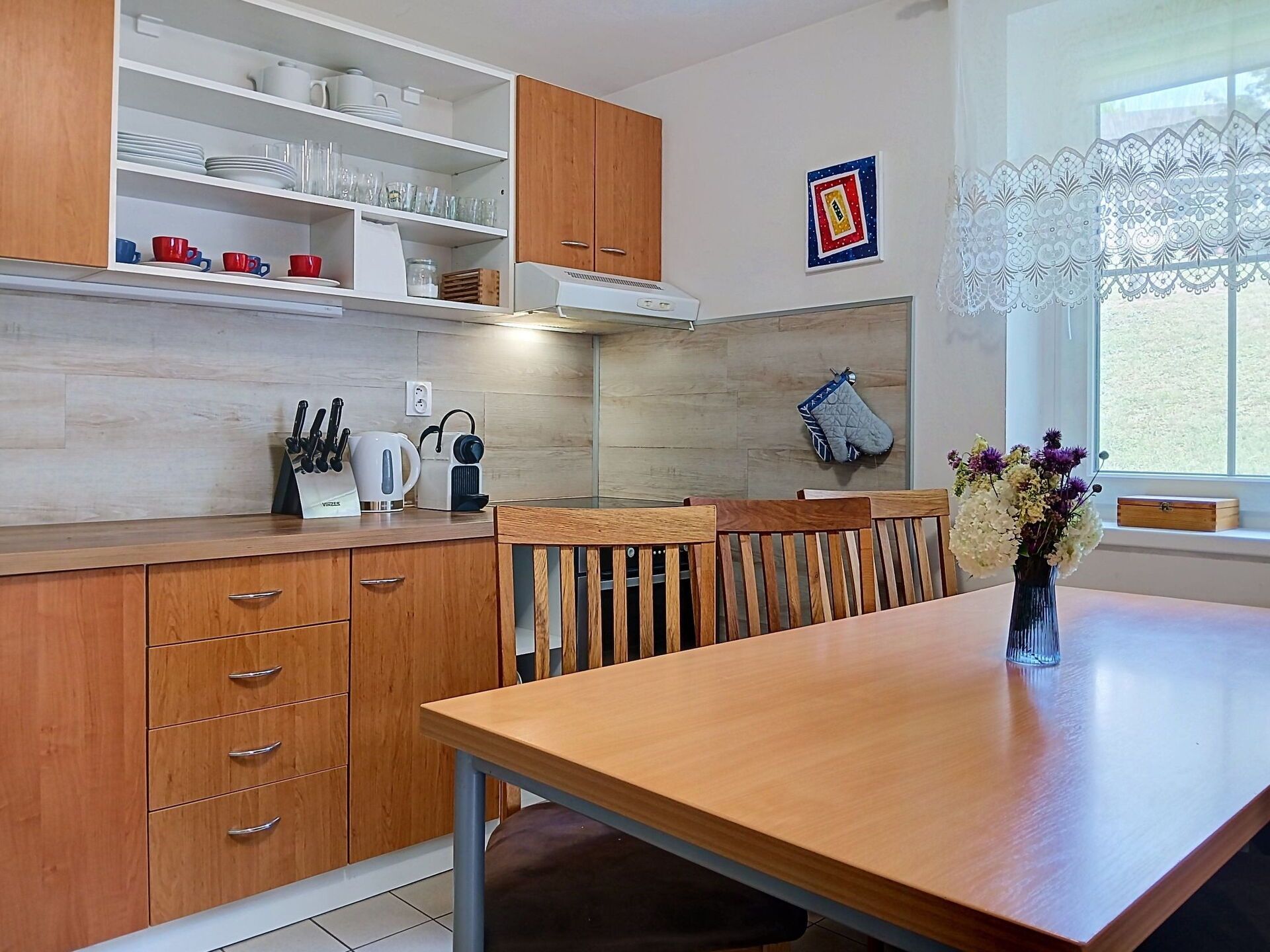 A modern kitchen with wooden cabinets, a dining table, and a window with lace curtains.