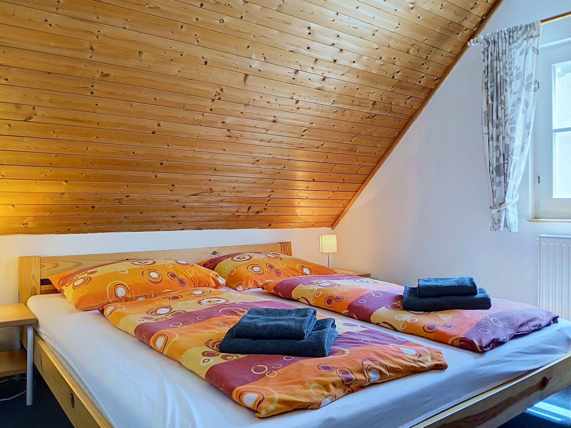 Een kamer met een schuine kap, een bed met kleurrijk beddengoed en een raam met gordijnen.