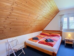 A bedroom with a sloped ceiling, a bed with colorful bedding, a bedside table with a lamp, and a chair.