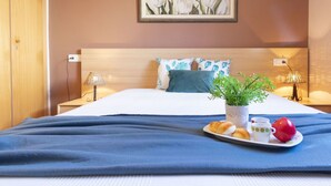 Un lit bien fait, recouvert d’une couverture bleue, une table de chevet avec une lampe, une œuvre d’art florale encadrée accrochée au mur, et une assiette contenant des croissants, une tasse et une pomme, posée sur le lit.