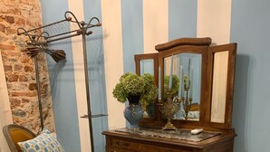 A room with a wooden chair with yellow upholstery, a blue patterned rug, a wooden desk with a mirror, and a vase with green plants.