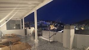 An outdoor terrace with wooden tables and chairs, patterned flooring, and a view of the city at night. - Muharrem Bey Butik Otel (Cesme)
