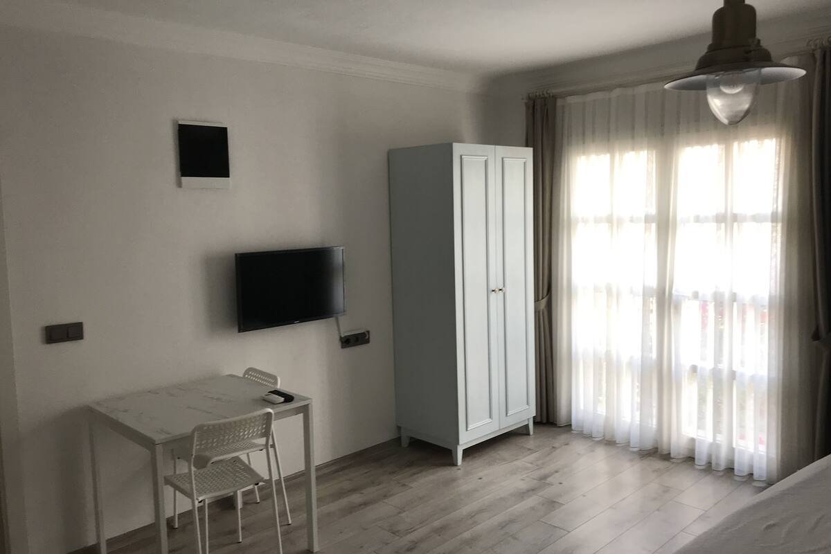 A room with a white desk, a television mounted on the wall, a wardrobe, and a window with curtains.