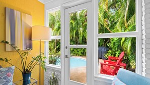 Um quarto claro com uma parede amarela, uma janela grande, uma piscina e uma cadeira vermelha lá fora.