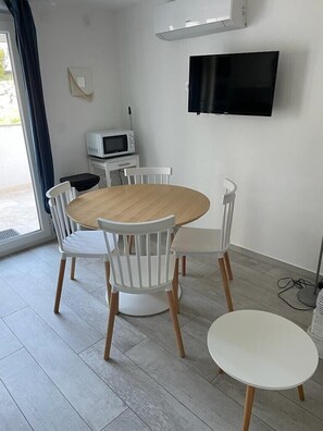 A dining area with a round wooden table and four white chairs, a small white table, a flat-screen TV mounted on the wall, and a microwave on a small white cabinet.