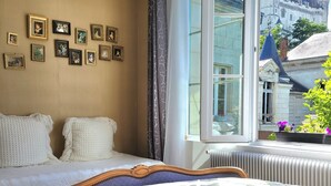 A bedroom with a large bed, a window with a view of a castle, and several framed pictures on the wall. - Splendid view of the Château and the Loire (Saumur)