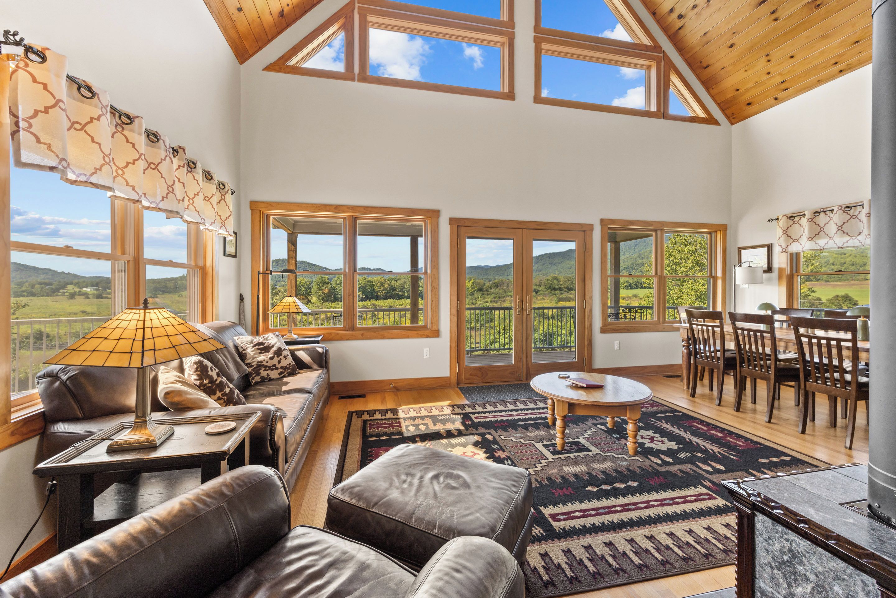 A spacious living room with a high ceiling, large windows, a leather sofa, a dining area, and a view of the countryside.