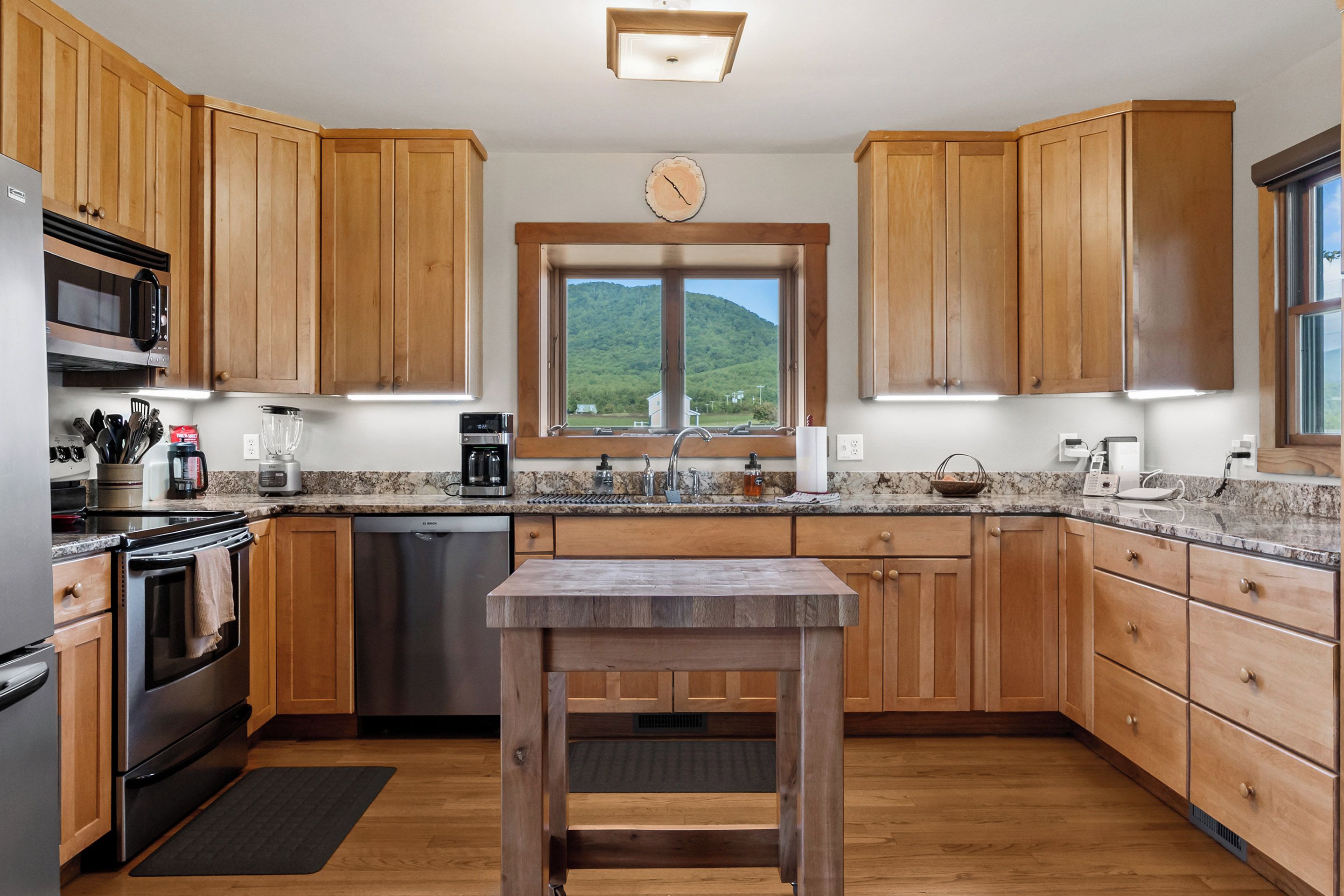 A modern kitchen with wooden cabinets, stainless steel appliances, a marble countertop, and a view of a green hillside through a window.