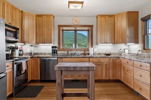 A modern kitchen with wooden cabinets, stainless steel appliances, a marble countertop, and a view of a green hillside through a window.