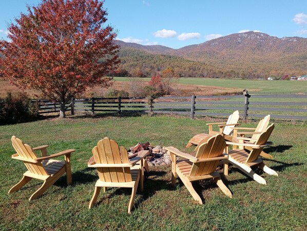 Un grupo de sillas Adirondack de madera dispuestas alrededor de un brasero en un espacio al aire libre con césped, con vistas a una cadena montañosa y una valla de madera al fondo.
