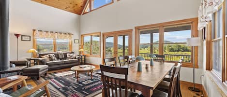 A spacious living area with a wooden ceiling, large windows, a sofa, a dining table, and a view of the outdoors.