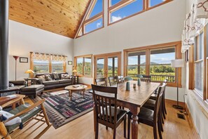 A spacious living area with a wooden ceiling, large windows, a sofa, a dining table, and a view of the outdoors.