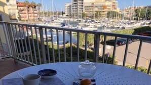 A round table with a white tablecloth, two white mugs, a glass jar, and a plate with a single orange on a balcony overlooking a marina with boats and buildings. - T2 comfortable Marina de Canet Plage (Canet-en-Roussillon)