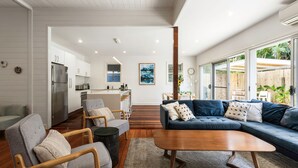 A modern living room with a blue sofa, wooden coffee table, and grey armchairs. A kitchen with white cabinets and a refrigerator is visible in the background. - Aaloka Bay - heritage charm w. pool & spa (Byron Bay)