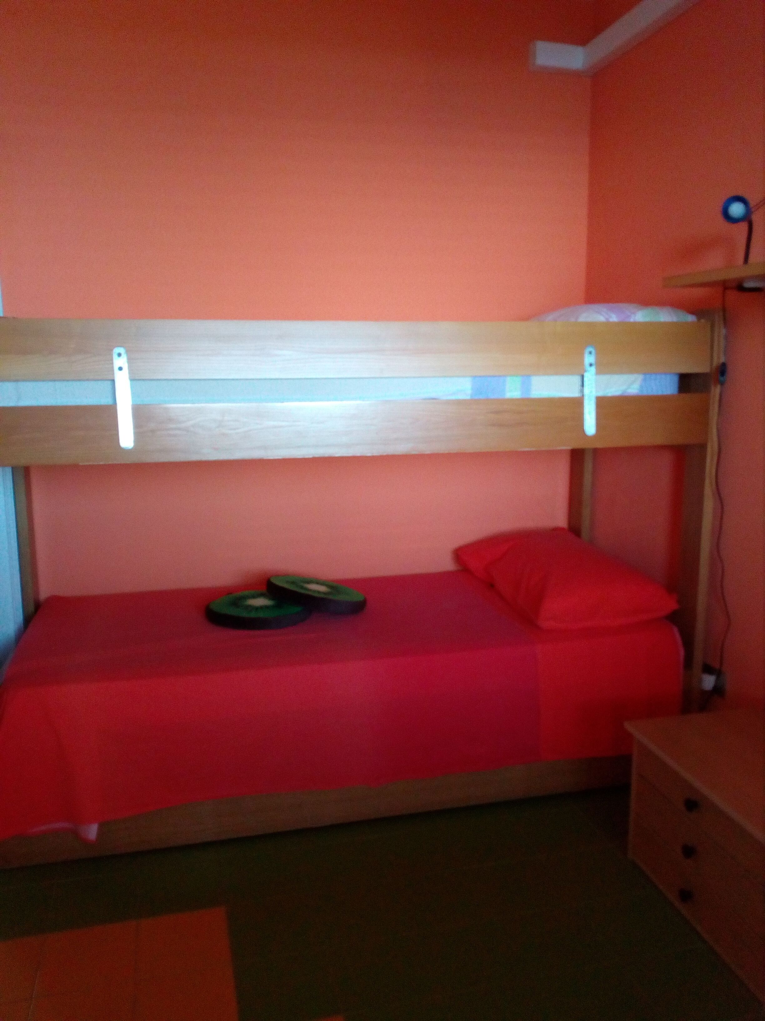 A bunk bed with a red mattress and pillows, a small wooden nightstand, and a wall-mounted lamp.