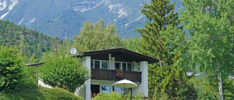 A two-story mountain cabin with a balcony, surrounded by lush greenery and mountains in the background.