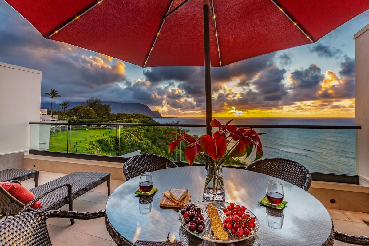 A round table with a glass top, surrounded by wicker chairs, a red umbrella, and a scenic view of the ocean and hills.