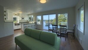 Ein geräumiger Wohnbereich mit einem grünen Sofa, eine Küche mit weißen Schränken und Blick auf einen See durch große Fenster.