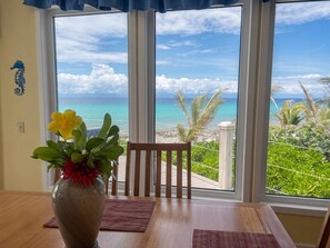 A room with a large window overlooking a beach, a wooden table, and a vase with flowers. - Second Wind - Ocean Front - 2 Bedroom (Utila)