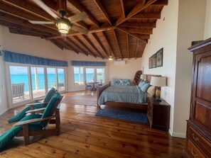 A bedroom with a wooden ceiling, a bed, a nightstand, a dresser, and a view of the ocean through large windows. - Second Wind - Ocean Front - 2 Bedroom (Utila)