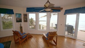 A room with wooden flooring, two wooden chairs, a ceiling fan, and large windows with blue curtains. - Second Wind - Ocean Front - 2 Bedroom (Utila)