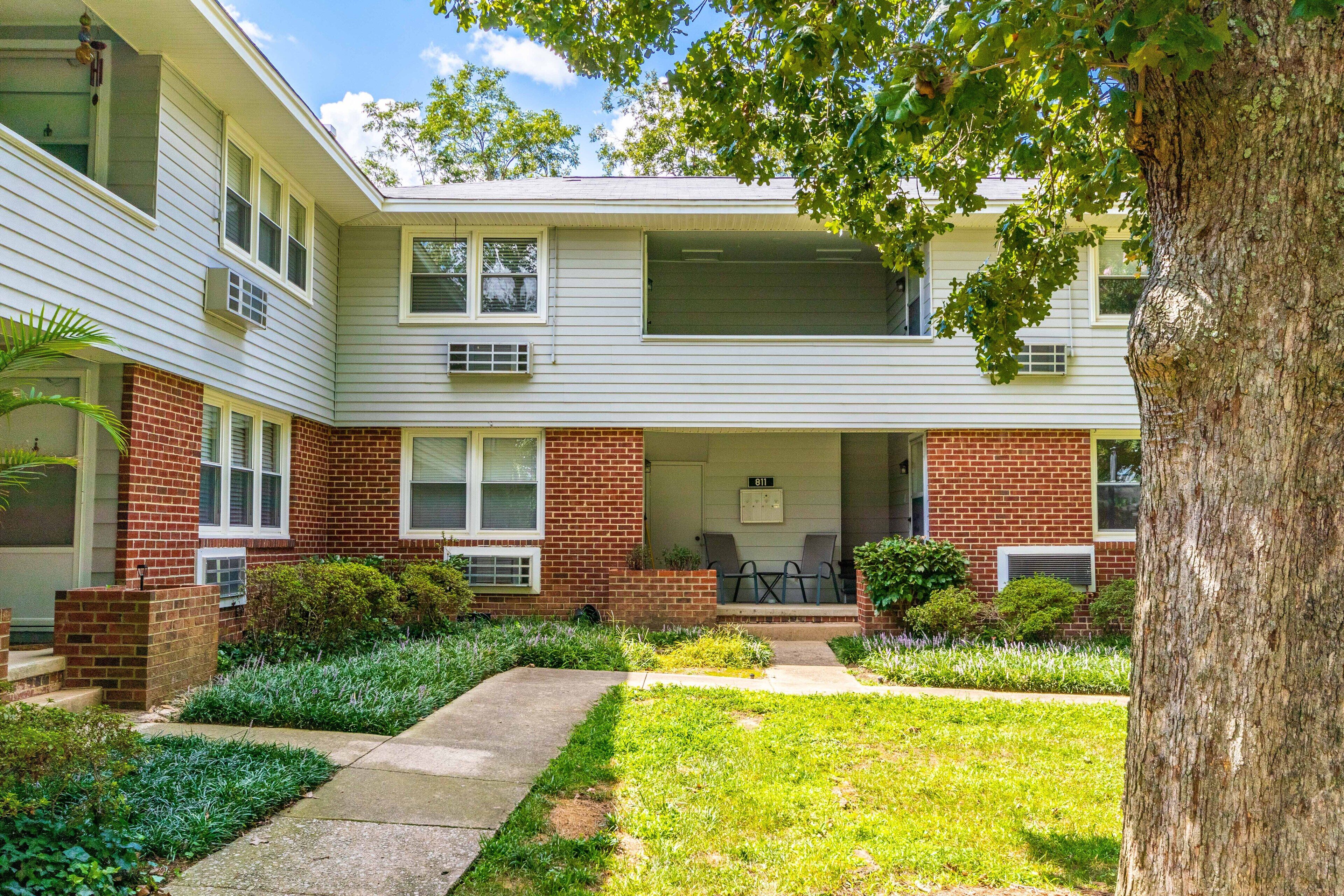 A two-story apartment building with a brick facade, a covered front porch with chairs, and a well-maintained lawn.