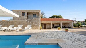 A stone building with a red-tiled roof, a swimming pool, and white lounge chairs.