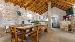 A rustic stone interior with a wooden dining table and benches, a kitchen in the background, and a stone fireplace.