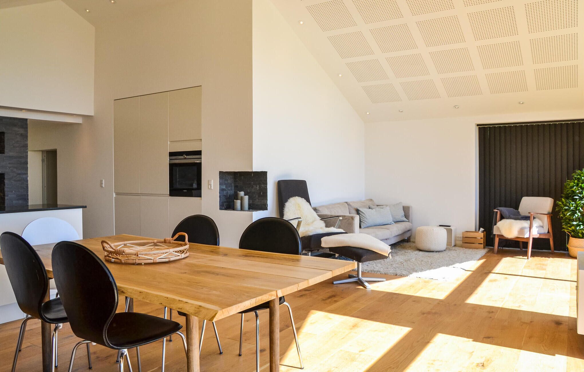 A modern living space with a wooden dining table, black chairs, a sofa, and a large window allowing natural light.