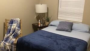 A bedroom with a bed, a bedside table with a lamp, a potted plant, and a chair with a blanket. - JLS Apartments Comples (Vero Beach)