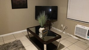 A living room with a flat-screen TV, a small table with a plant, a framed picture on the wall, and a window with blinds. - JLS Apartments Comples (Vero Beach)