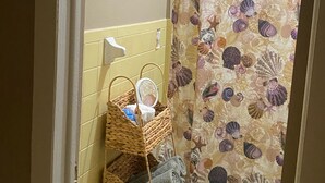 A bathroom with a yellow tiled wall, a shower curtain with a seashell pattern, a wicker basket with towels, and a framed picture above the mirror. - JLS Apartments Comples (Vero Beach)