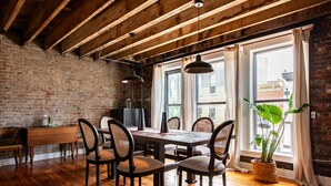 A dining area with a wooden table and chairs, a brick wall, and a large window with curtains. - Quaint Historic Downtown: Walk to Dinner & Shops, Poker Table, Rooftop, Garage (New Albany)