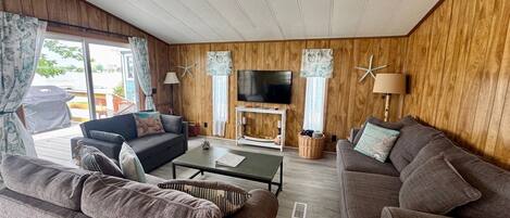 A cozy living room with two sofas, a coffee table, and a TV. The room has wooden walls and large windows.