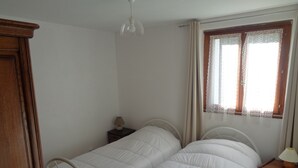 Un dormitorio con dos camas individuales, un armario de madera, una mesita de noche con lámpara y una ventana con cortinas de encaje.