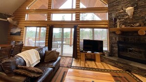 A log cabin interior with a large wooden ceiling, a stone fireplace, a leather sofa, and a television.