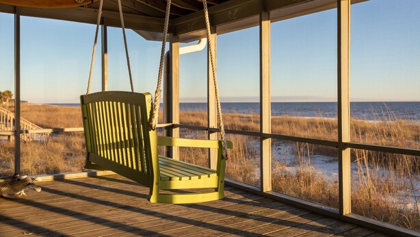 Eine Holzterrasse mit einem grünen Schaukelstuhl mit Blick auf einen Strand mit hohem Gras und dem Meer.