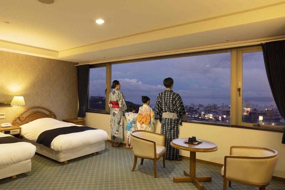 A hotel room with two beds, a sitting area, and a large window with a city view.