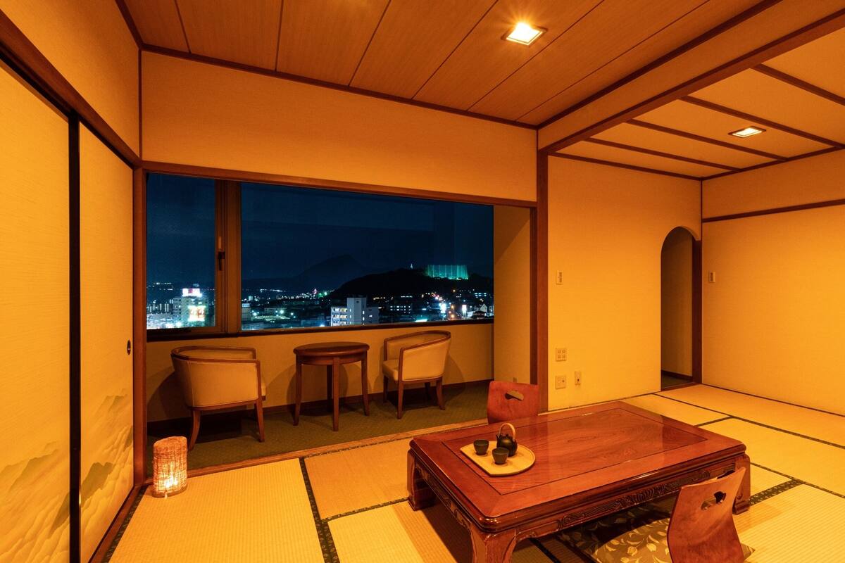 A traditional Japanese room with tatami flooring, a wooden table with tea set, and a view of a cityscape at night.