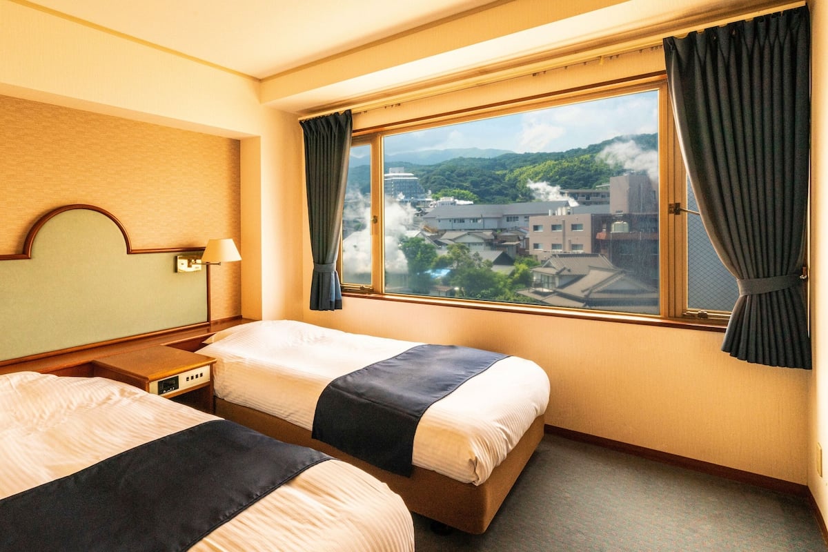 Habitación de hotel con dos camas, vista a un paisaje urbano y una ventana con cortinas.