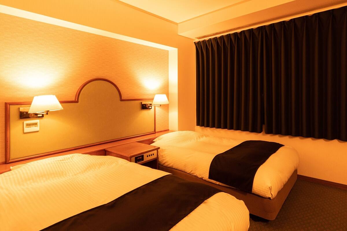 A hotel room with two beds, a headboard with built-in lamps, a control panel, and a large window with curtains.